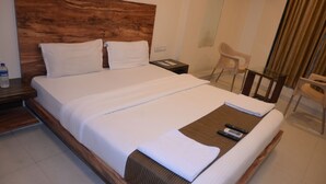 Una cama bien hecha con cabecera de madera, una mesita de noche con un teléfono y un control remoto, y una silla en una habitación de hotel.
