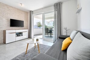 A modern living room with a flat-screen TV, a grey sofa with a yellow pillow, a white cabinet, and a glass door leading to an outdoor area. - One-bedroom Apartment with Balcony - C1 Infinity (Novalja)