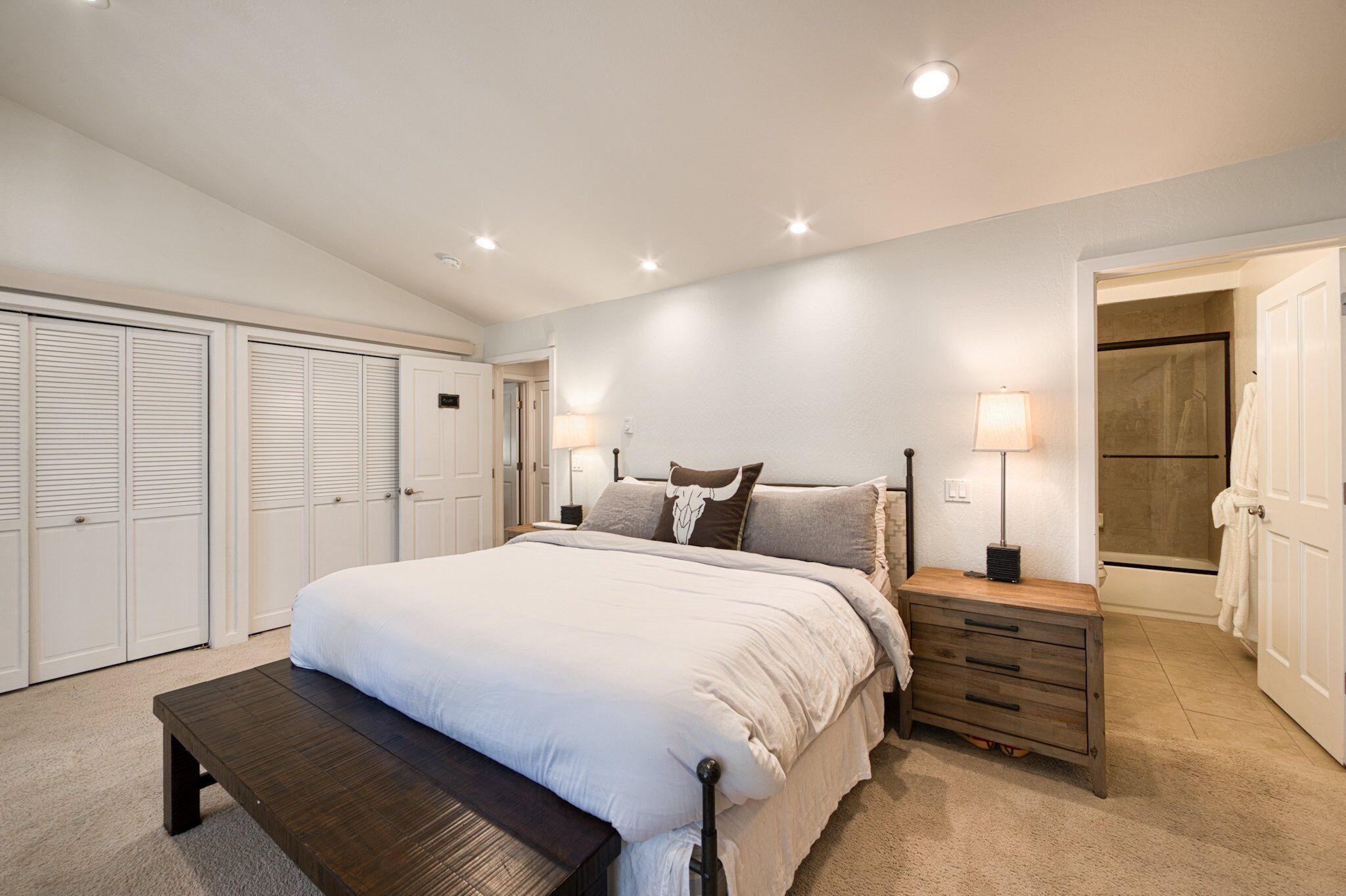 A bedroom with a bed, bedside tables, a bench, a walk-in closet, and a bathroom with a shower and towel rack.