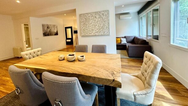 Un vaste espace de vie comprenant une table à manger en bois, des chaises à revêtement gris, un canapé beige et un grand tableau abstrait accroché au mur.
