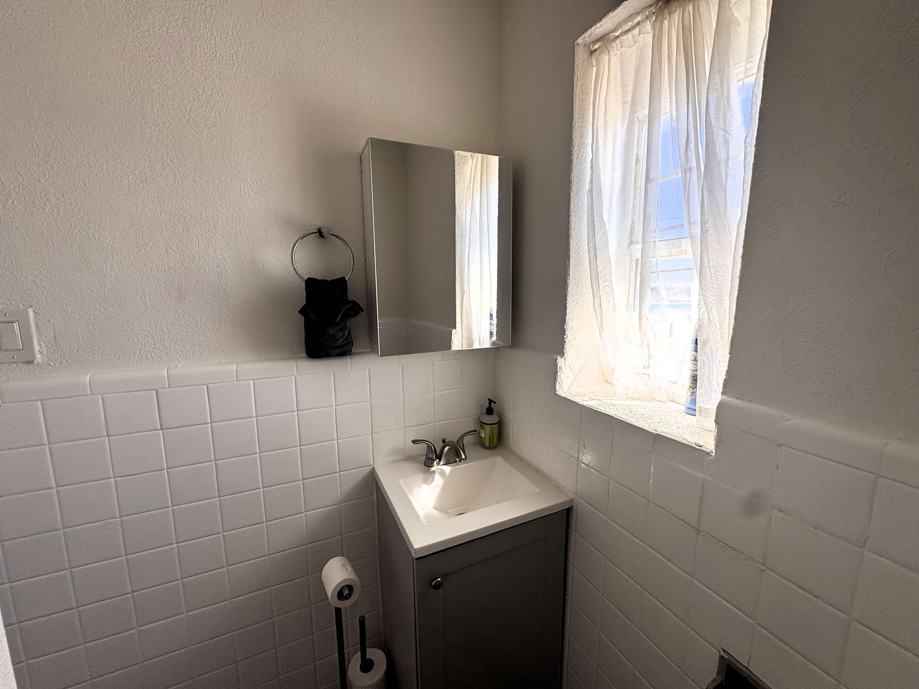 A bathroom with white tiled walls, a sink with a mirror above it, a window with sheer curtains, and a toilet paper holder.