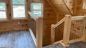 Eine Holzhütte mit Schlafzimmer im Obergeschoss, einem Fenster mit Blick auf Bäume und einem Holzgeländer.