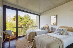 Ein Schlafzimmer mit zwei Betten, einem Fenster mit Blick auf Bäume, einem Korbstuhl und Holzboden.