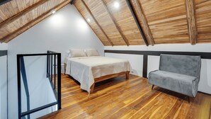 Camera soppalco con soffitto spiovente, pavimento in legno, un letto, un divano grigio e una ringhiera in vetro.