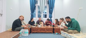 A group of people seated around a wooden table in a room with a window and curtains.