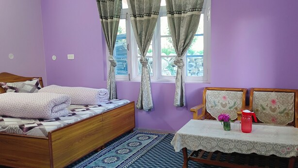 Un dormitorio con una cama, una mesa con un florero y una termo, y una ventana con cortinas.
