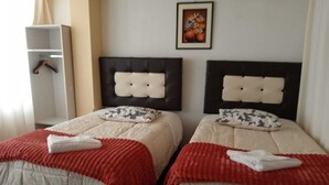 Habitación de hotel con dos camas, cada una con cabecero capitonado, mesita de noche blanca y colcha con estampado rojo y blanco.