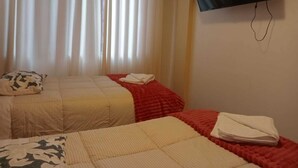 Una habitación con dos camas, cada una con un edredón de estampado diferente, una manta roja y un televisor montado en la pared.