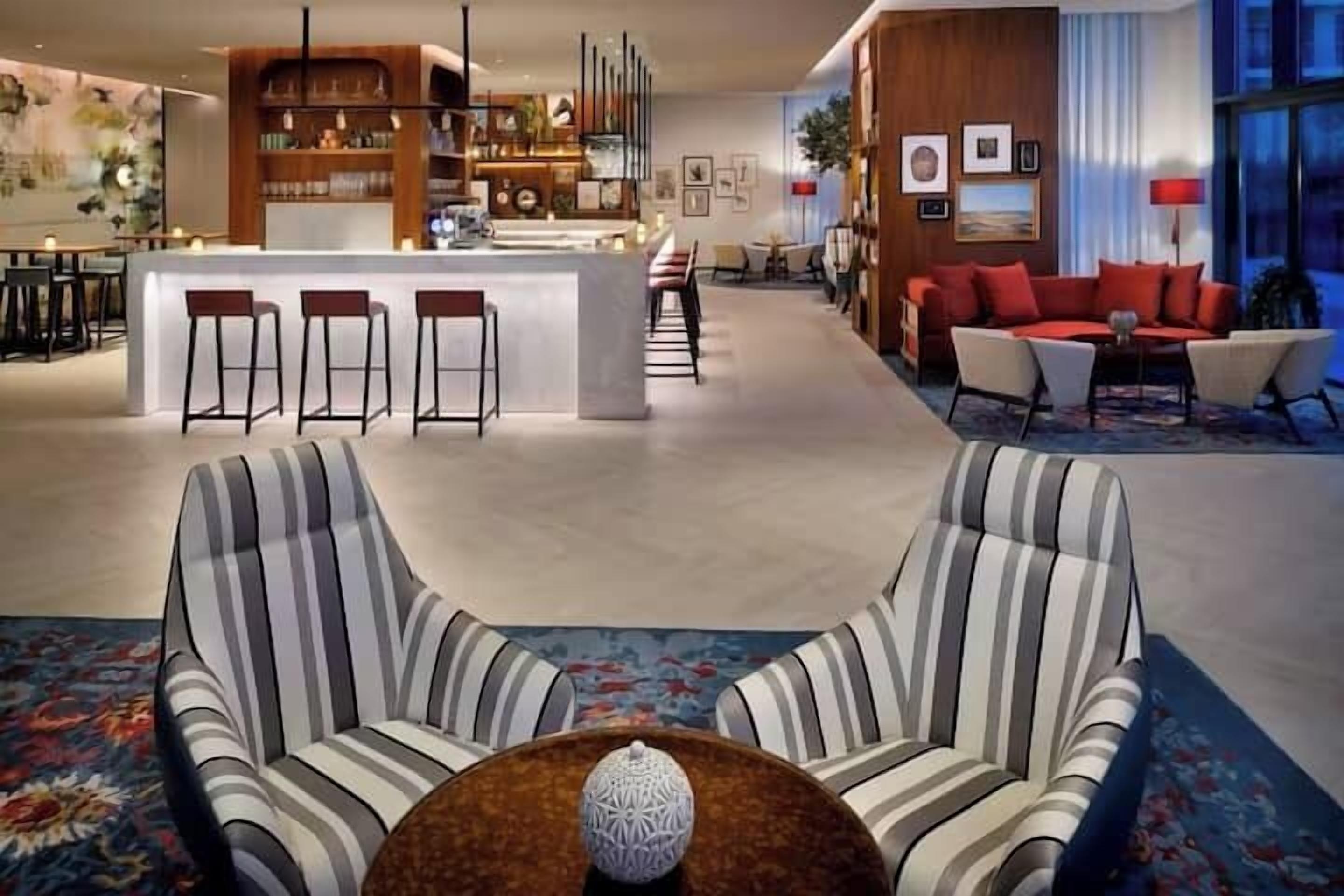 A modern hotel lobby with striped sofas, a bar area, and a seating area with red couches.