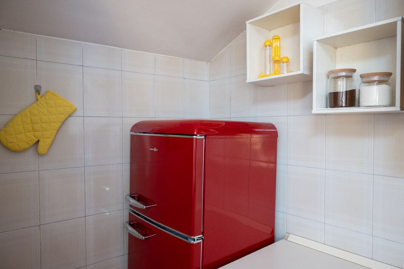 Un vieux réfrigérateur rouge dans une cuisine aux murs recouverts de carreaux blancs, un manique jaune et des étagères remplies de bocaux.