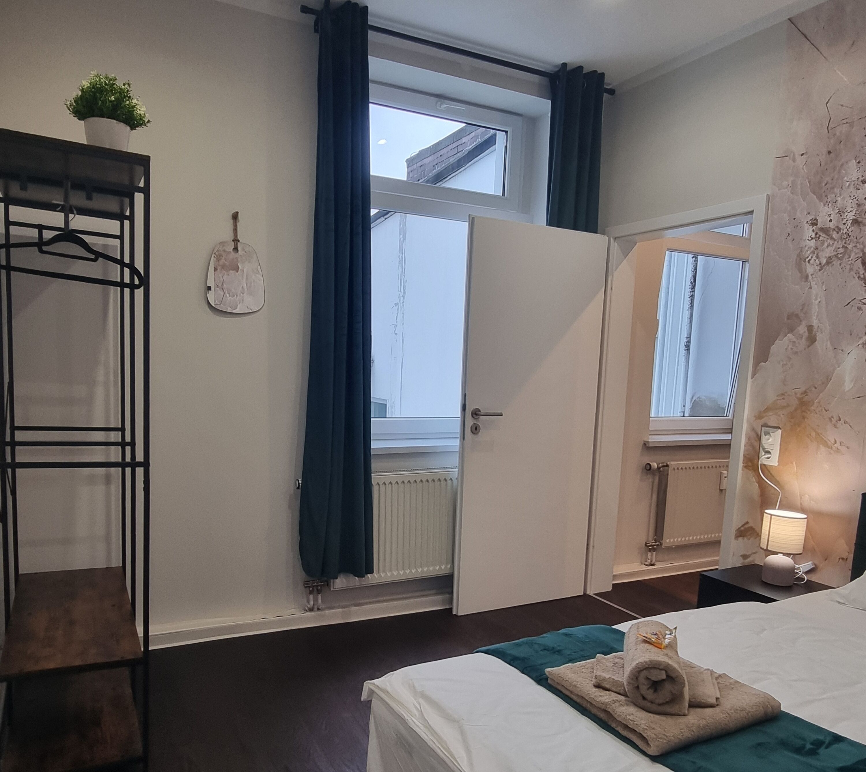 A bedroom with a bed, a radiator, a window with curtains, a mirror, a towel, and a shelf with a plant.