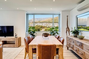 A modern dining area with a large window offering a city view, a wooden dining table, leather chairs, a TV on a wooden stand, and a giraffe sculpture. - 2bed Mouille Point Apartment w Amazing Cape Views (Cape Town)