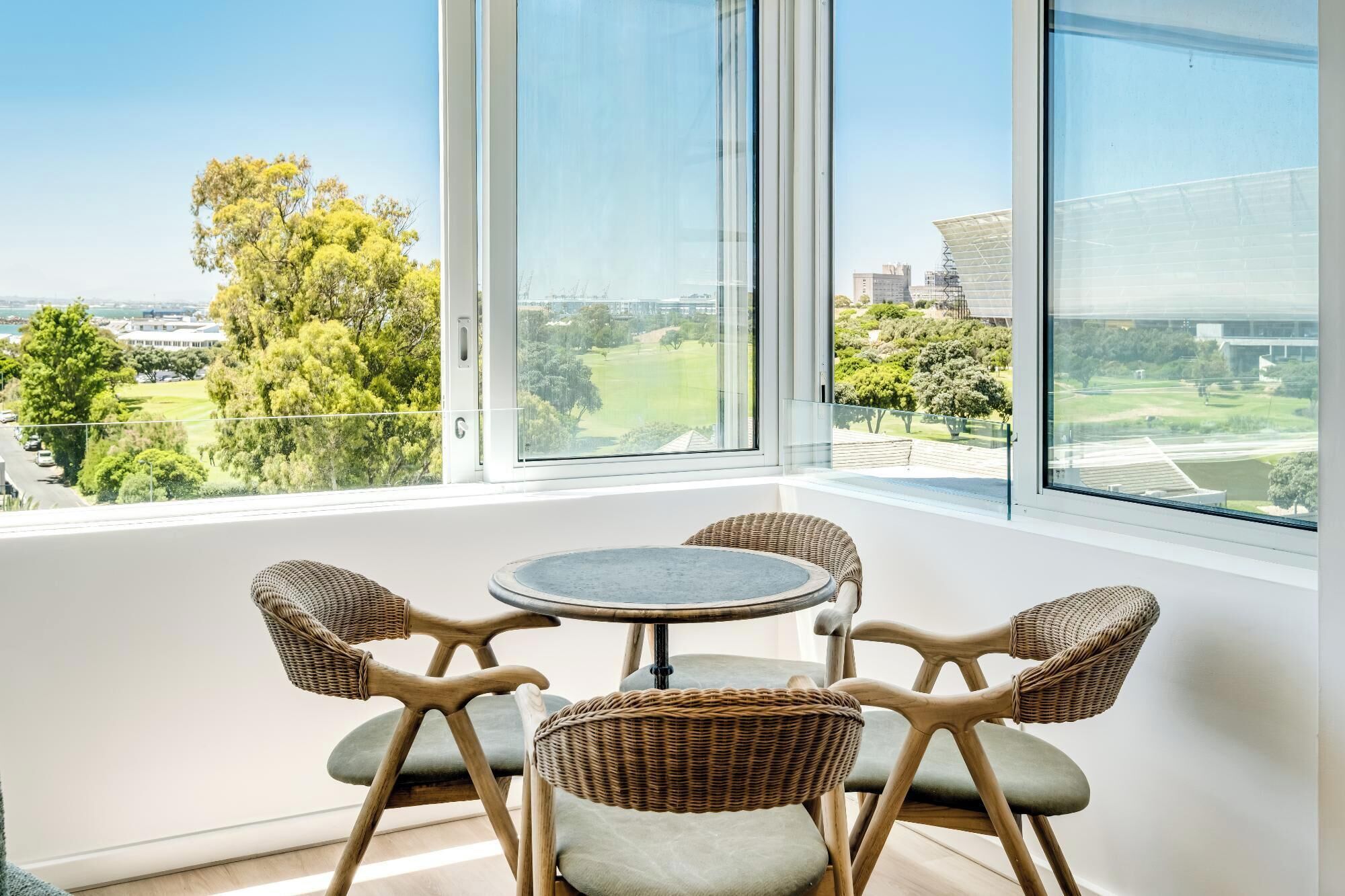 A balcony with a round table and four wicker chairs, overlooking a green landscape and buildings.