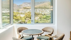 Ein Zimmer mit großem Fenster, das auf eine Stadtlandschaft und Berge blickt. Vor dem Fenster steht ein runder Tisch mit zwei Stühlen.