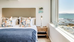 Ein Schlafzimmer mit Bett, Kissen, einem Nachttisch mit Lampe und Blick aufs Meer.