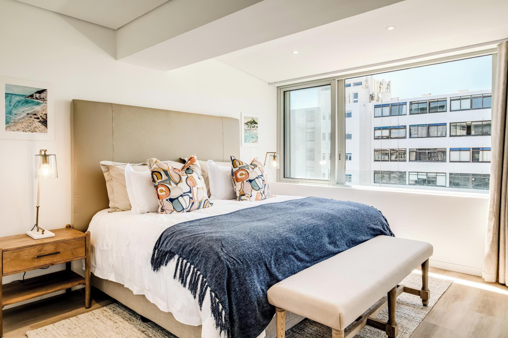 Une chambre moderne avec un grand lit, un banc, une table de chevet et une vue sur un immeuble par la fenêtre.