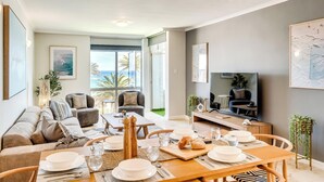 A modern living room with a dining area, large windows with a view of the ocean, and a flat-screen TV. - Camps Bay Seaside Beach View Apartment Palm Bay (Cape Town)