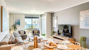 A modern living room with a dining area, large windows with a view of the ocean, and a flat-screen TV.
