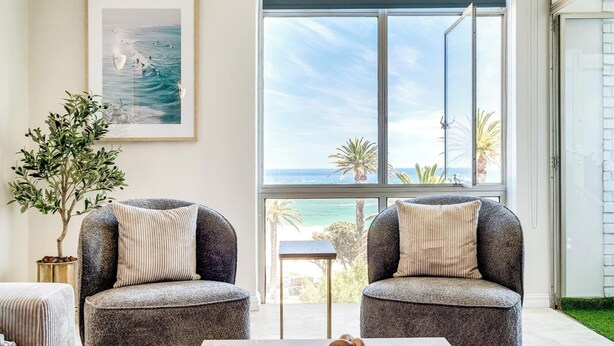 A room with two grey armchairs, a small table with a decorative object, a framed picture on the wall, and a large window with a view of the sea and palm trees. - Camps Bay Seaside Beach View Apartment Palm Bay (Cape Town)