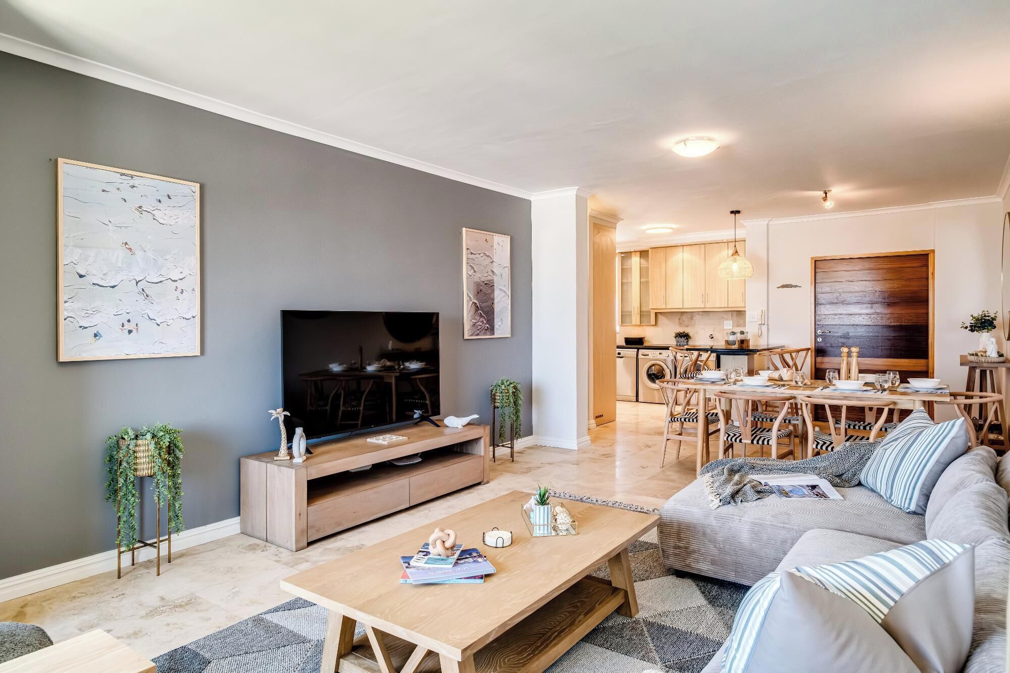 Een moderne woonkamer met een flatscreen televisie, een houten salontafel en een bank met gestreepte kussens. Er is een eethoek met een houten tafel en stoelen, en een keuken op de achtergrond.