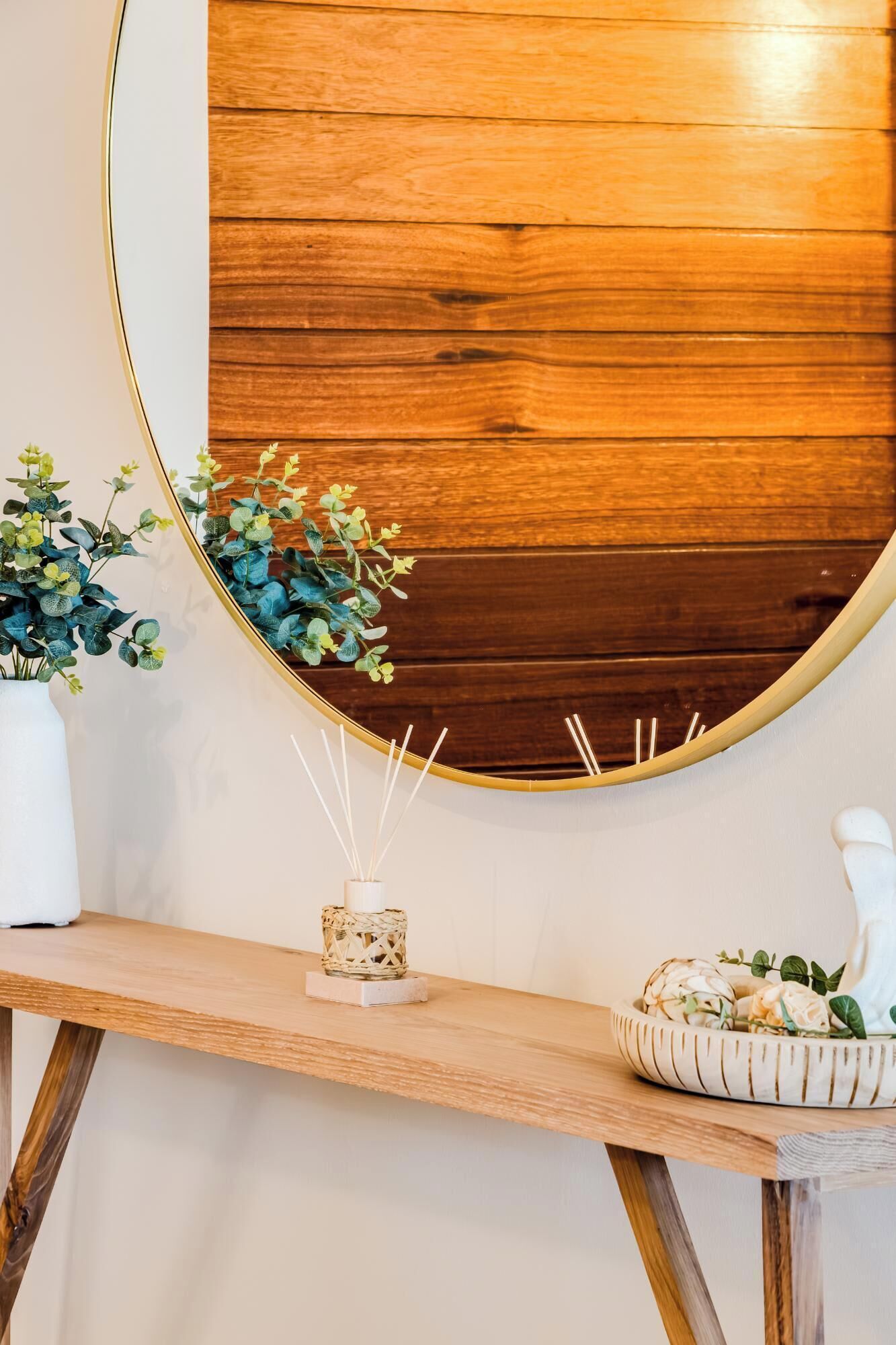 Un mur en bois orné d’un miroir rond, un vase blanc contenant des plantes vertes, un petit porte-bougie en osier et un bol décoratif rempli de fleurs.