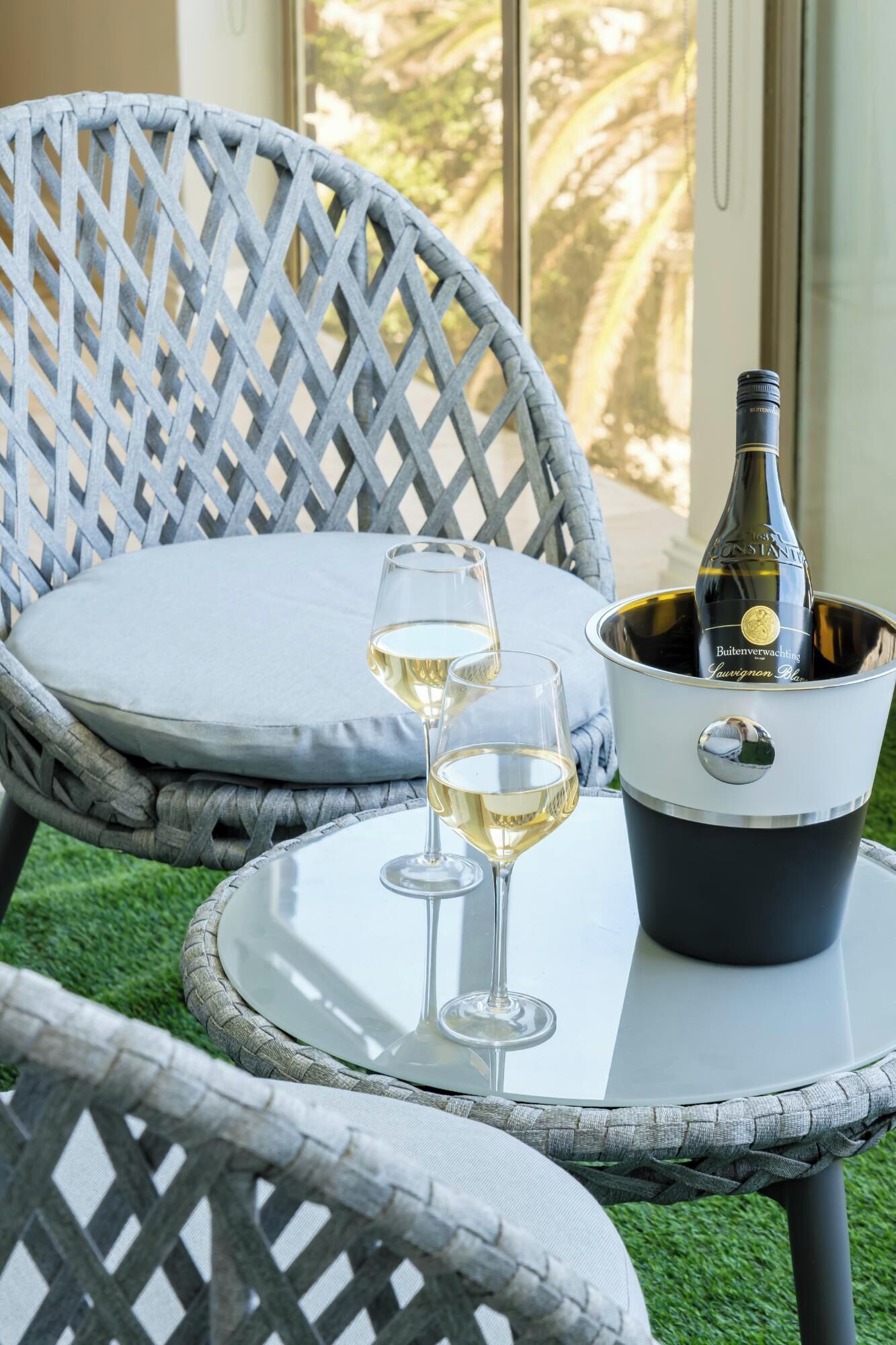 A wicker chair with a cushion, a glass table, two wine glasses, and a bottle of wine in a bucket.