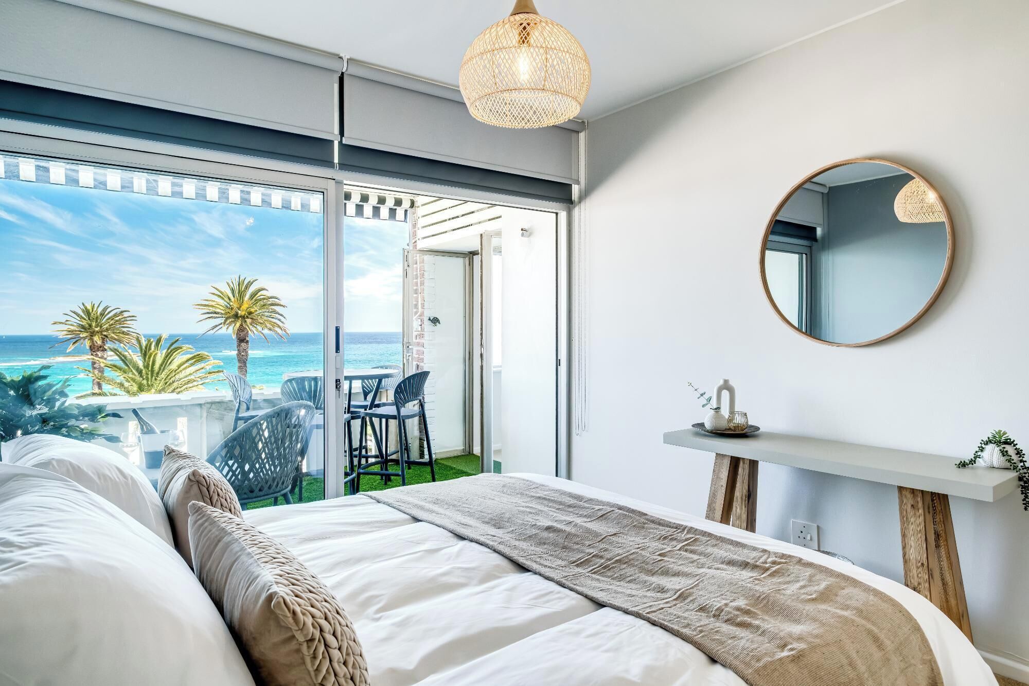A bedroom with a large bed, a view of the ocean, a balcony with chairs, and a round mirror on the wall.