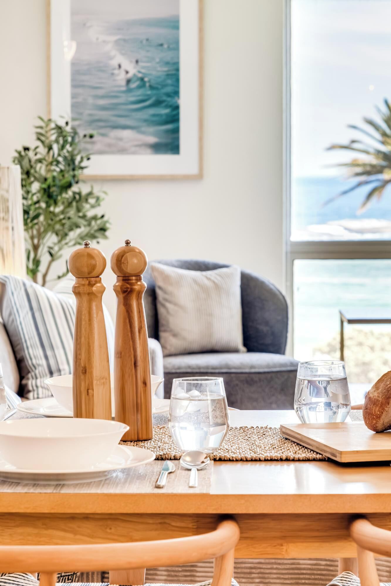 A dining table set with plates, glasses, and a bread roll, with a view of the ocean through a window.