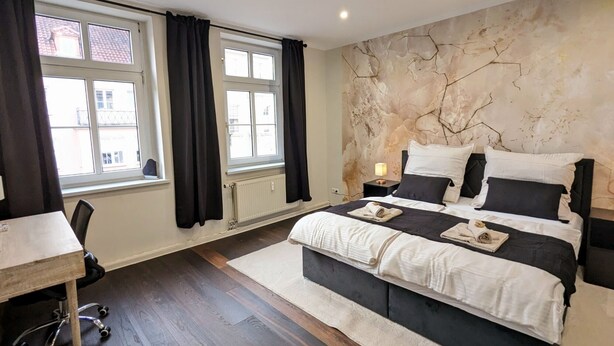 Ein Schlafzimmer mit einem großen Bett, einem Schreibtisch und einem Stuhl. Zwei Fenster mit Vorhängen, ein Heizkörper und eine Wand mit strukturierter Gestaltung sind zu sehen.