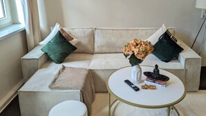 Ein Wohnzimmer mit einem beigefarbenen Sofa, einem runden Couchtisch und einem kleinen weißen Tisch. Über dem Sofa hängen drei gerahmte Kunstwerke an der Wand.