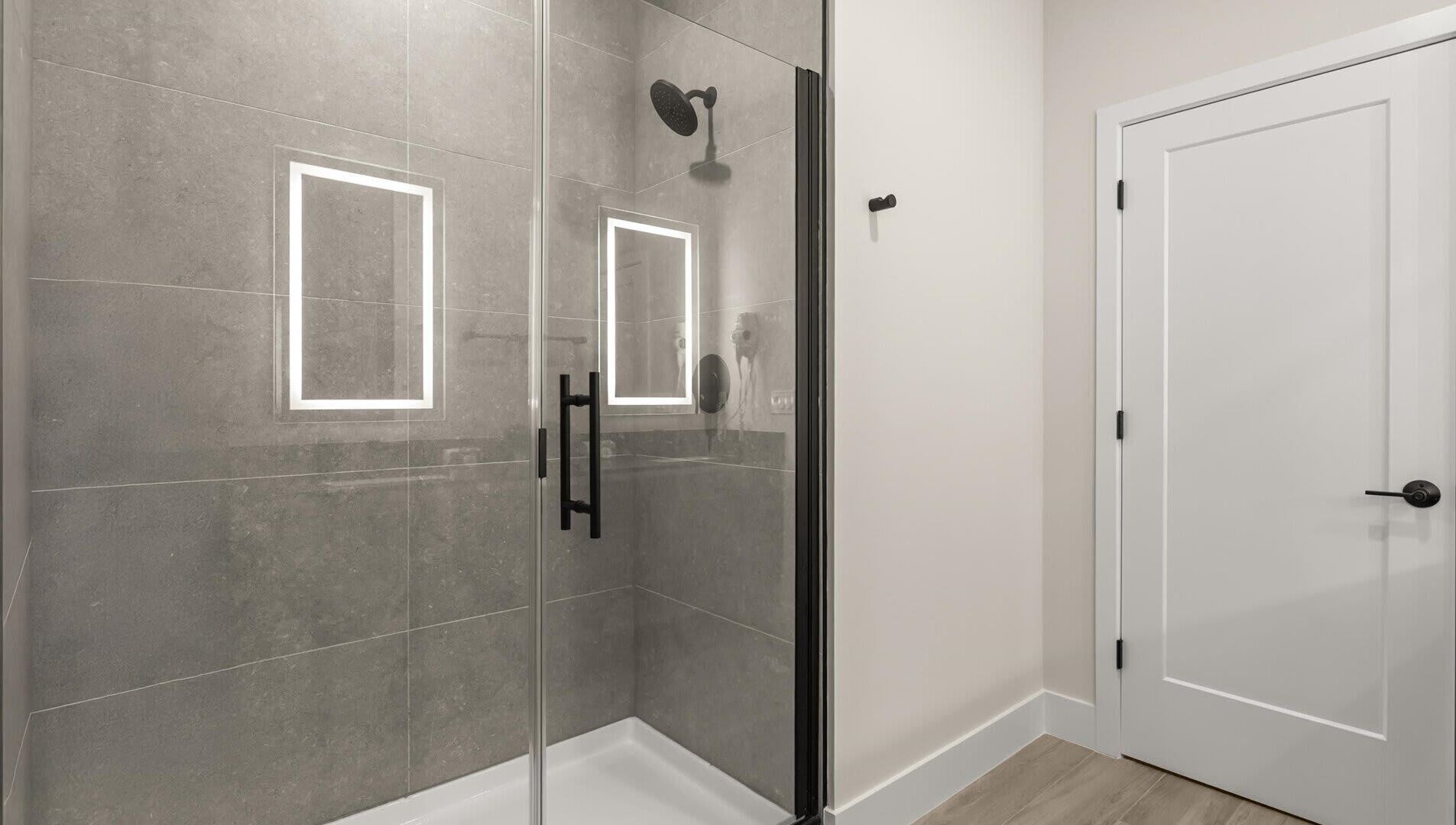 A modern bathroom with a glass-enclosed shower, a white door, and grey tiled walls.