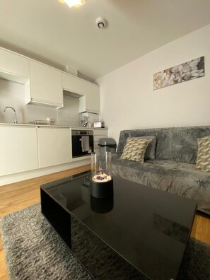 A modern living room with a black glass coffee table, grey sofa, and a kitchen area with white cabinets and a stove. - Luxury home from home 2 (Kent)