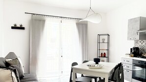 Una sala de estar moderna y luminosa con un sofá, una mesa de comedor y sillas. Hay una zona de cocina con una cafetera y un estante con objetos decorativos.
