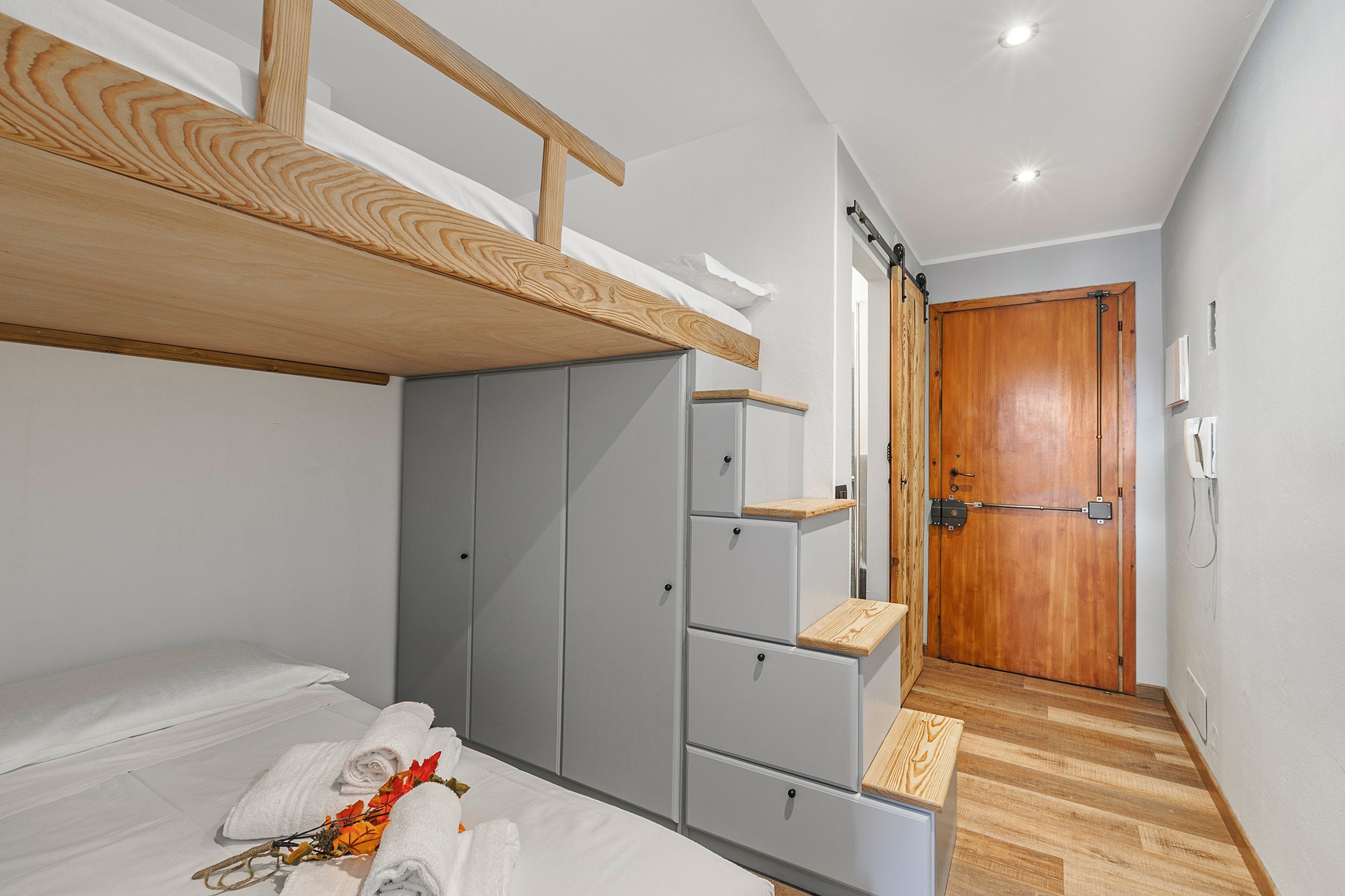 Un dormitorio con una cama litera, un armario gris, una puerta de madera y un estante de madera pequeño.