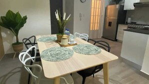 Een eethoek met een houten tafel, witte stoelen en groene placemats. Links staat een potplant en in de achtergrond is een keuken te zien.
