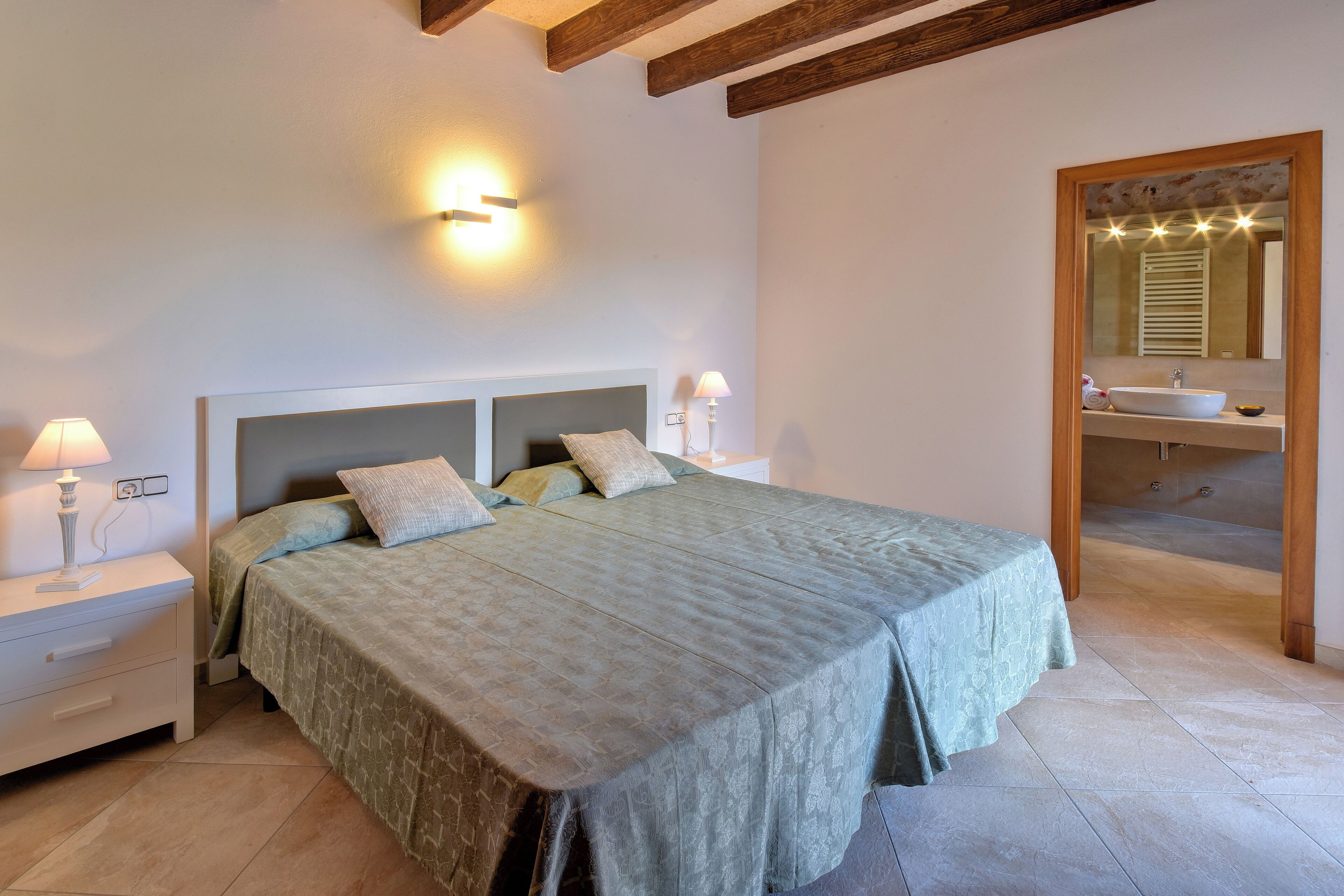 Een slaapkamer met een groot bed, bedlampen, een nachtkastje, een houten plafond en een badkamer met een wastafel en spiegel.