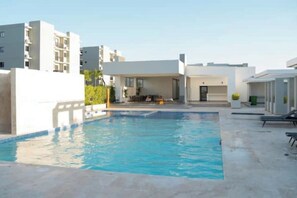 Un área de piscina moderna con una piscina azul cristalina, rodeada de edificios blancos y una zona de estar cubierta.