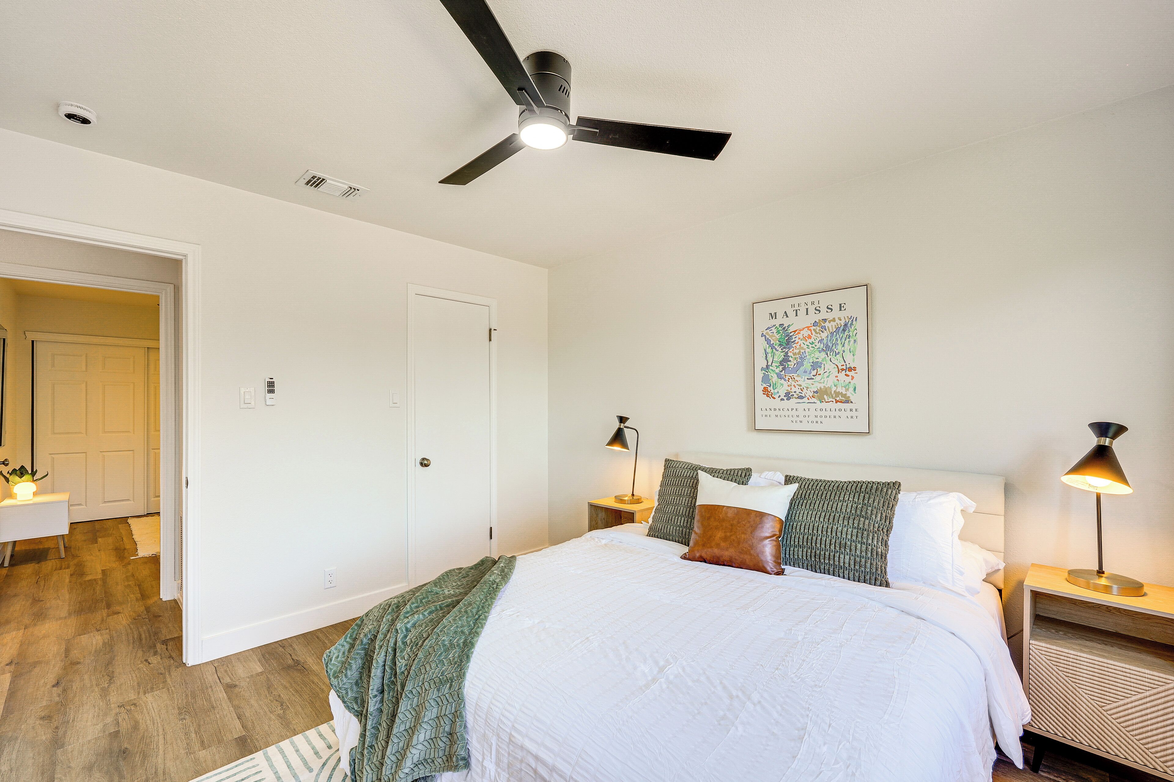 A bedroom with a bed, a ceiling fan, a nightstand with a lamp, a door, and a wall with a framed picture.