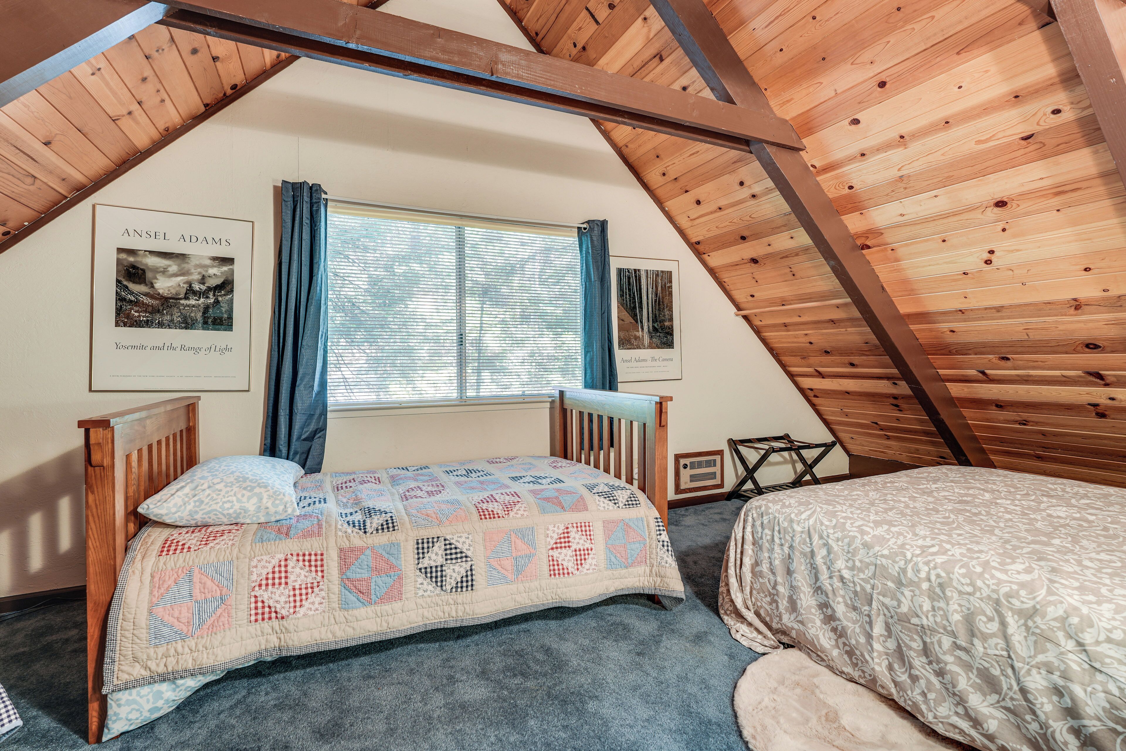 Un dormitorio acogedor en el ático, con techo inclinado, una cama con colcha, una ventana con cortinas y dos fotografías enmarcadas en la pared.