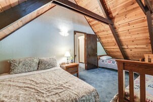 Un dormitorio con techo inclinado, una cama con ropa de cama estampada, una mesita de noche con lámpara y una puerta que da a otra habitación.