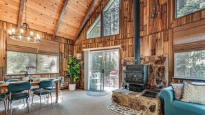 A cozy cabin interior with a dining area, a stone fireplace, and large windows offering views of the forest. - 4 Mi to State Park: Quiet Escape w/ Deck in Arnold (Arnold)