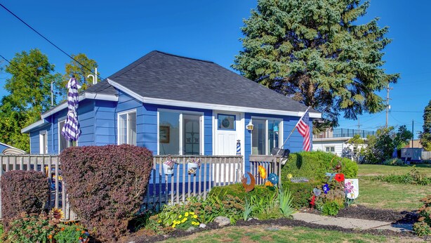 Una casa azul con valla de piquetes blancos, un pequeño jardín con diversas plantas y flores, y una bandera estadounidense.