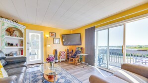 Una sala de estar con sofá, sillón y una mesa de centro. Hay un televisor montado en la pared y un ventanal con vistas.