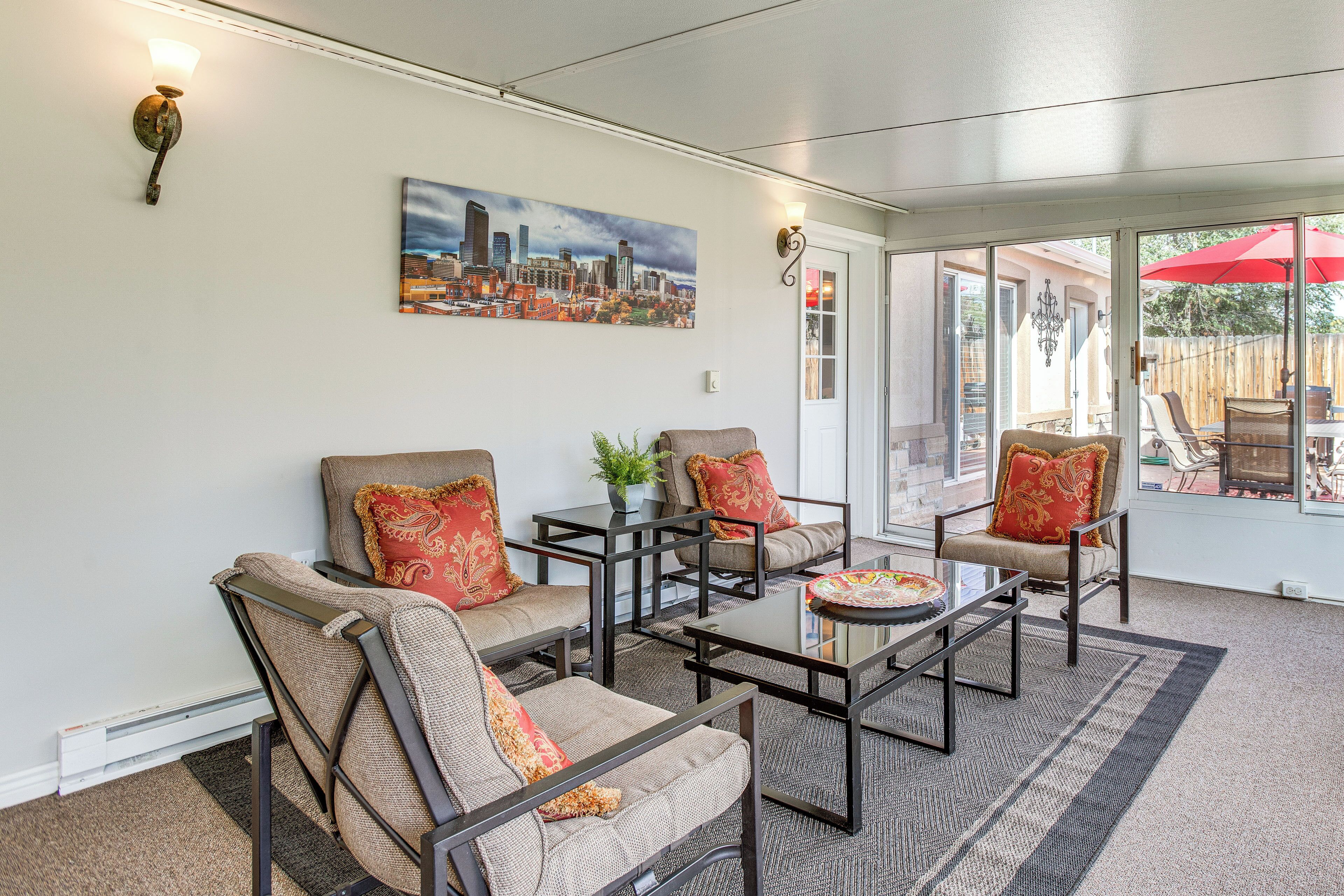 A sunlit room with a cityscape painting, a glass door leading outside, and a red umbrella visible through the glass.