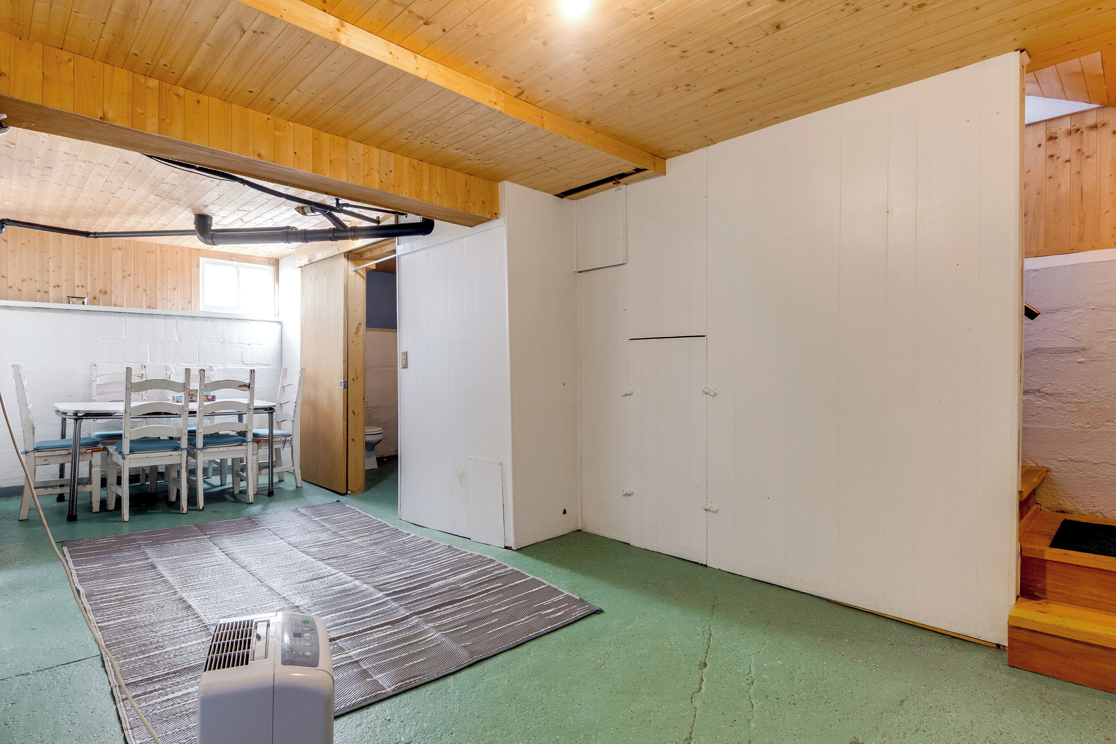 A room with a wooden ceiling, a green floor, a white wardrobe, and a table with chairs.