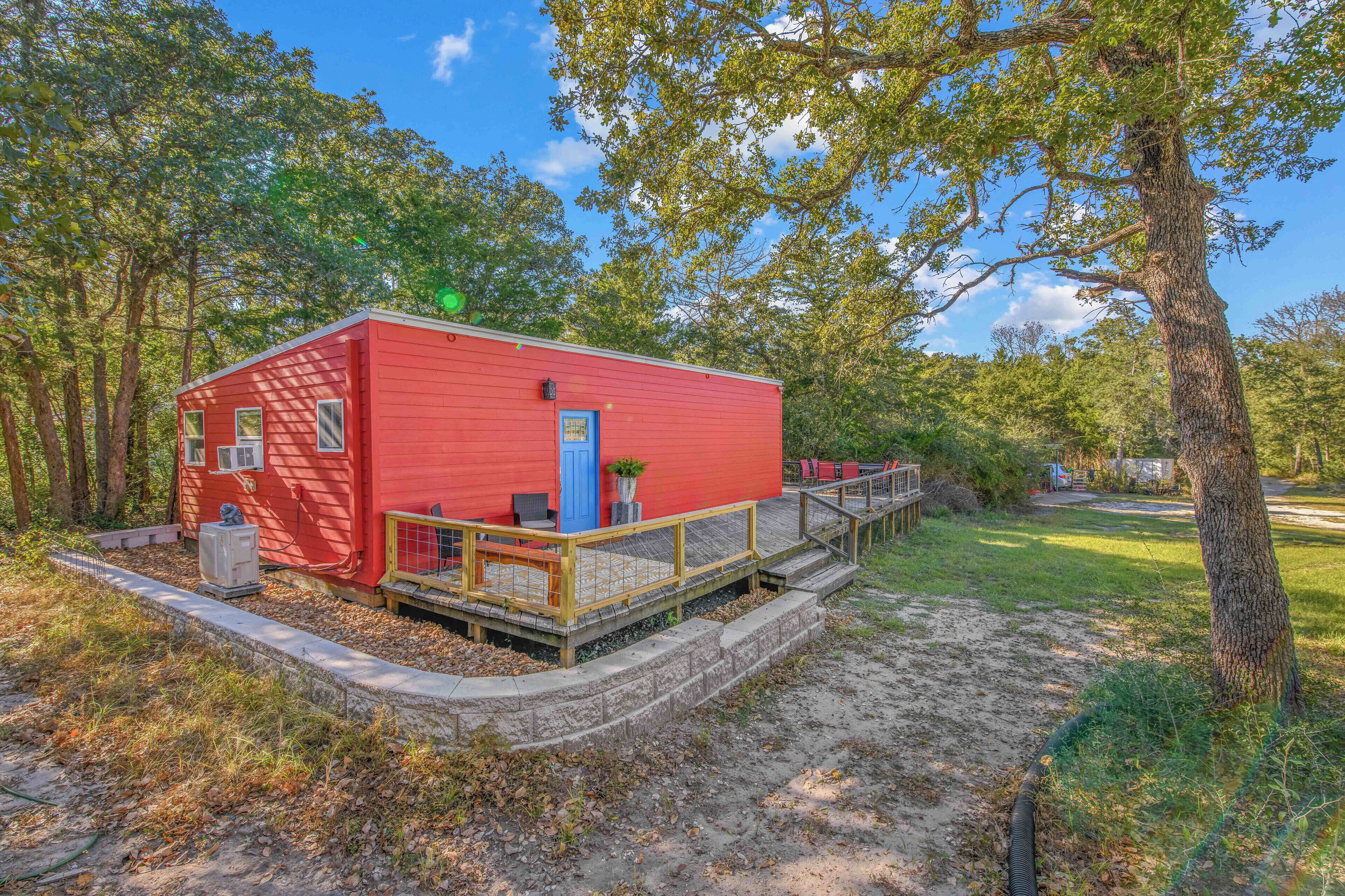 A red cabin with a wooden deck and a blue door, surrounded by trees and a clear sky.