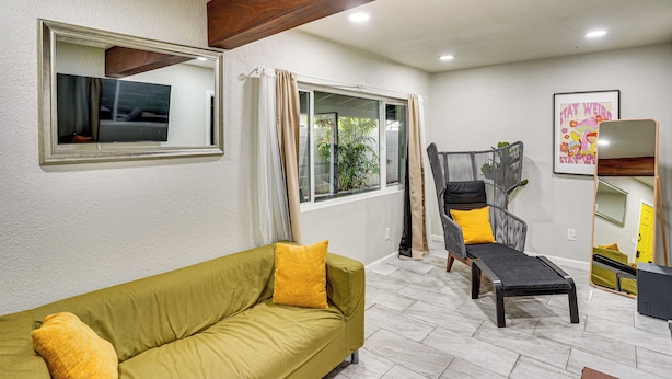 A living room with a green sofa, a black coffee table, a grey chair with yellow cushions, a wooden beam ceiling, and a large mirror. - Explore LA & Long Beach: Compton Condo w/ Patio (Compton)