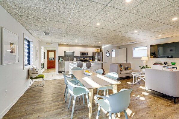 A modern kitchen with a central island, a dining area with a long table and chairs, and a living area with a sofa and TV.