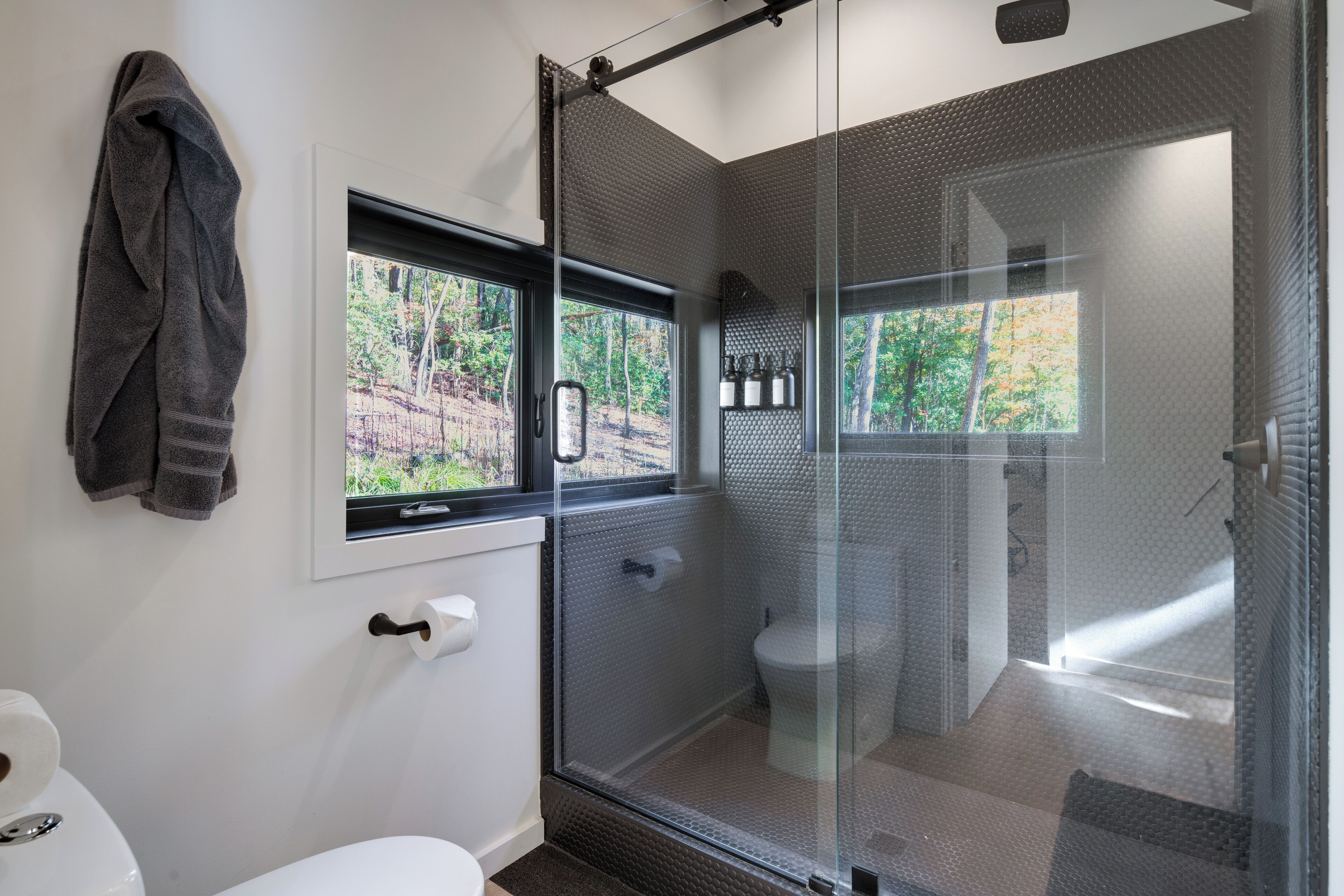 Un baño moderno con ducha envuelta en vidrio, inodoro y ventana con vistas a árboles.