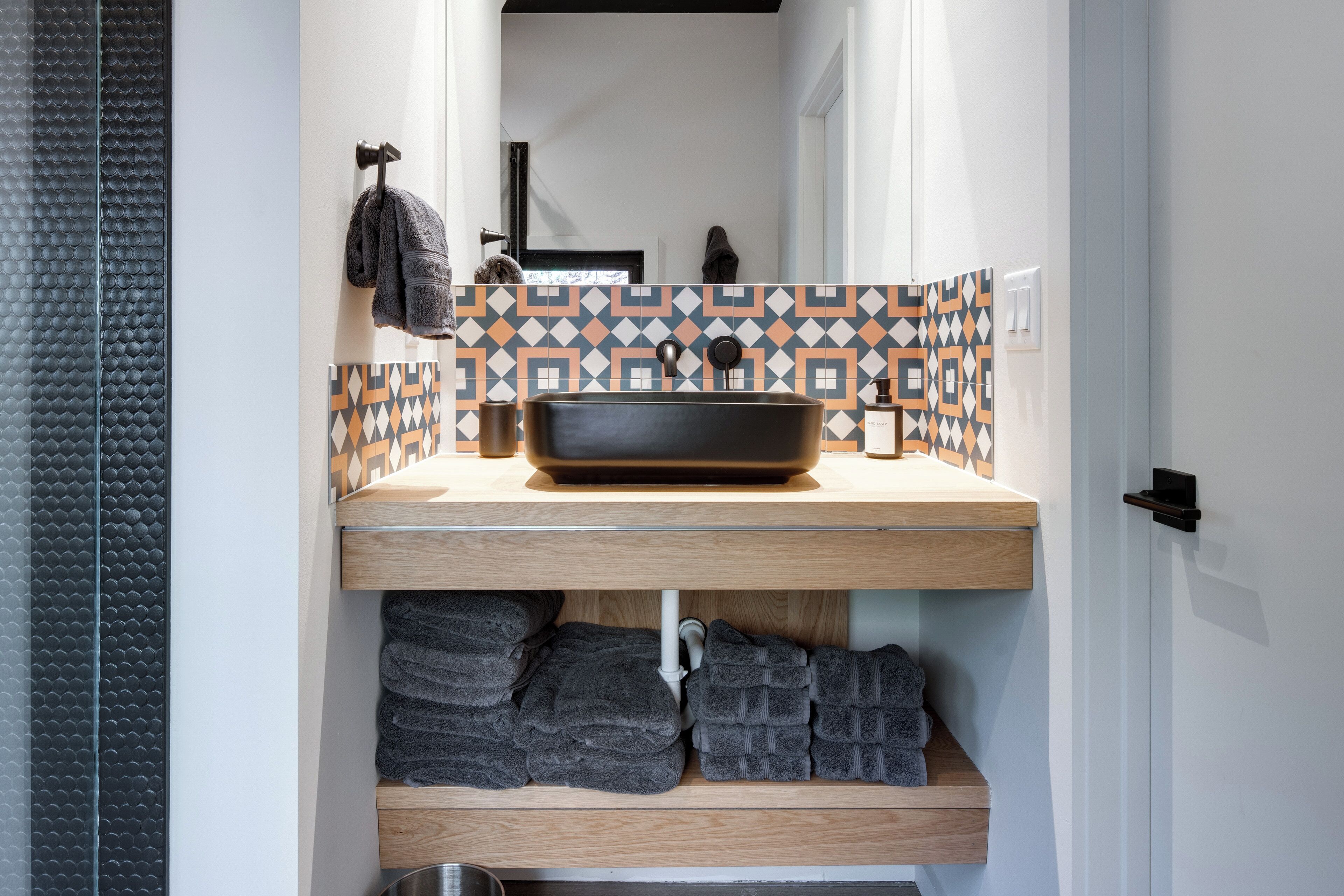 A bathroom with a black basin, patterned tiles, and neatly folded towels on shelves.